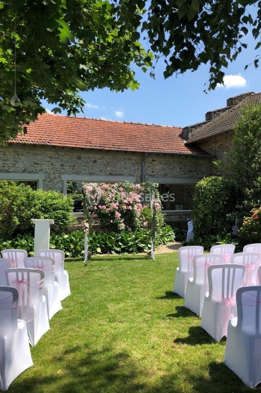 Chaises blanches décorées de rubans roses alignées devant une arche fleurie dans un jardin ensoleillé.