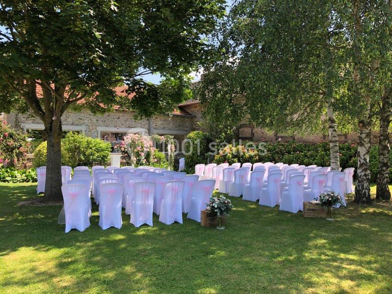 Chaises blanches alignées en extérieur, sous des arbres, préparées pour une cérémonie dans un jardin fleuri.