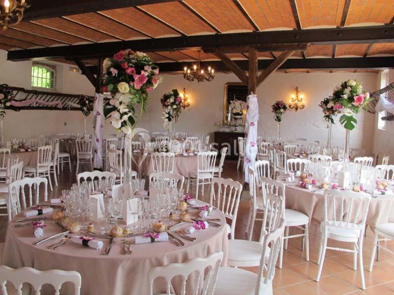 Salle de réception décorée avec des tables rondes, nappes beiges, chaises blanches et arrangements floraux colorés.