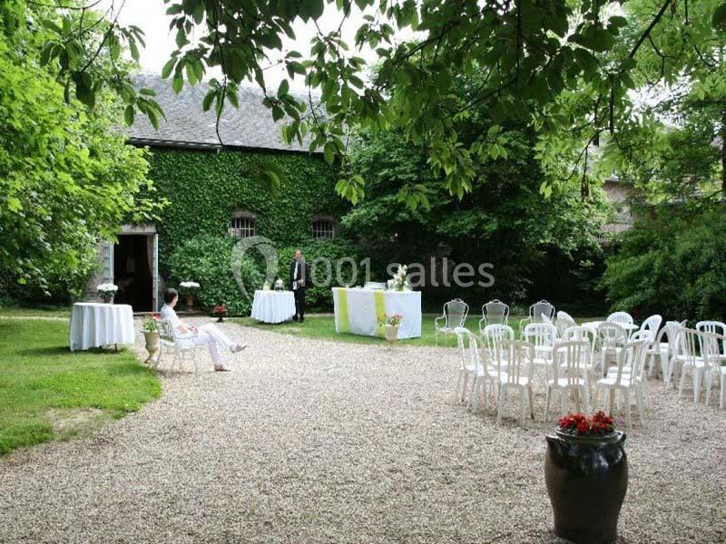 Cour extérieure avec chaises blanches disposées, tables décorées et bâtiment en pierre couvert de lierre en arrière-plan.
