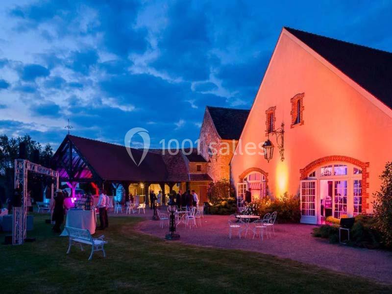 Bâtiment éclairé en soirée avec des invités rassemblés à l'extérieur, entourés de mobilier de jardin.
