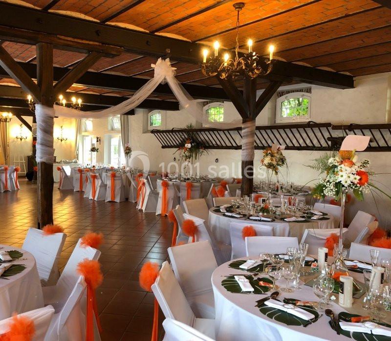 Salle de réception décorée avec des tables rondes, nappes blanches, chaises ornées de nœuds orange et arrangements floraux.