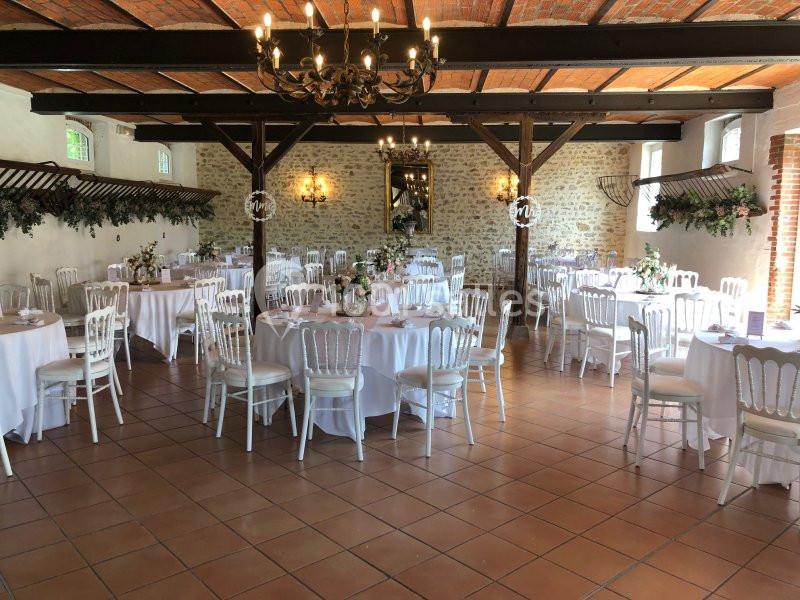 Salle de réception décorée avec des tables rondes dressées, des chaises blanches et des éléments floraux sur les murs.