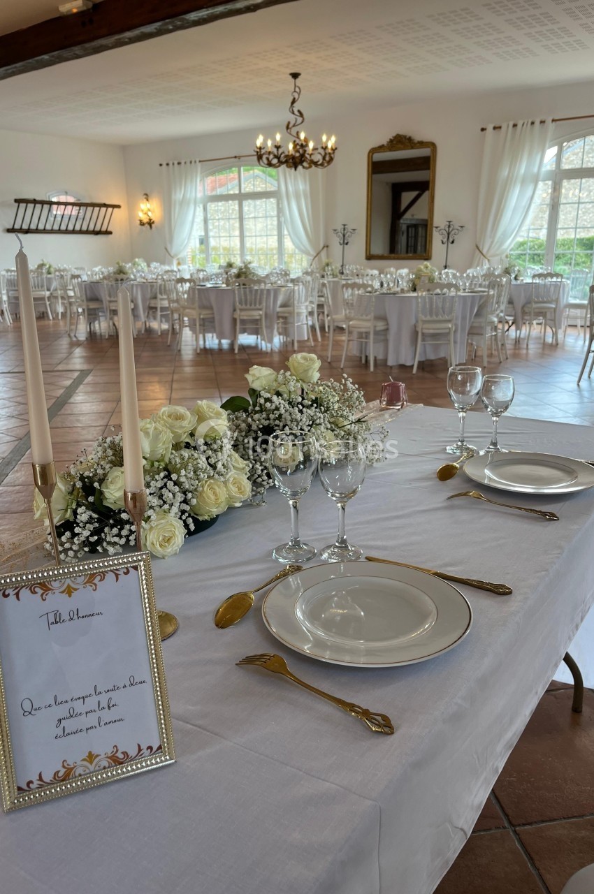 Salle de réception élégante avec tables dressées, décorations florales blanches et chandeliers, vue sur des fenêtres…