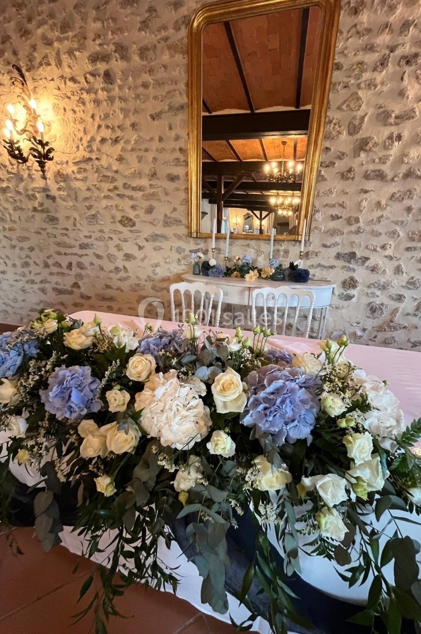 Décoration florale avec roses blanches et hortensias bleus sur une table, devant un mur en pierre et un miroir doré.