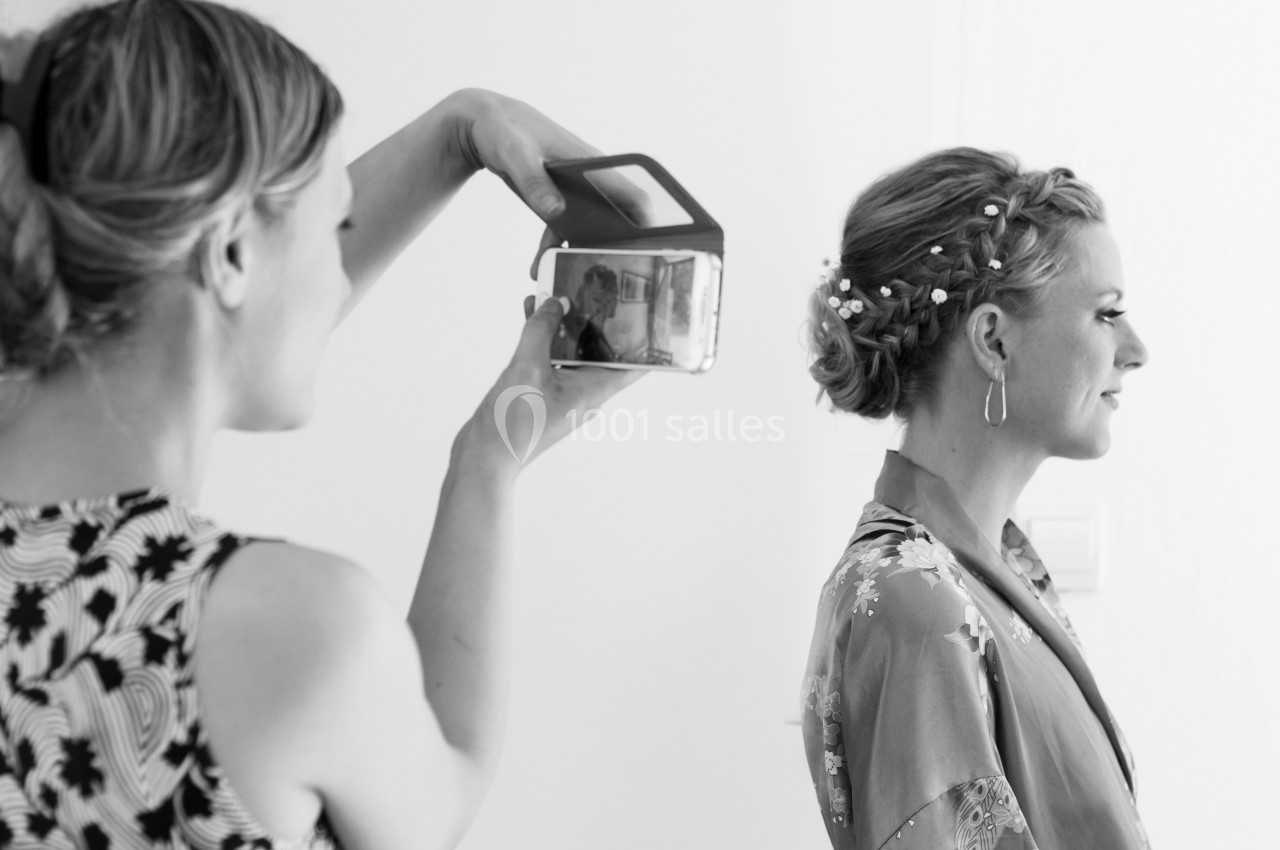 Une femme prend une photo d'une autre femme coiffée et maquillée, portant un peignoir, dans un intérieur lumineux.