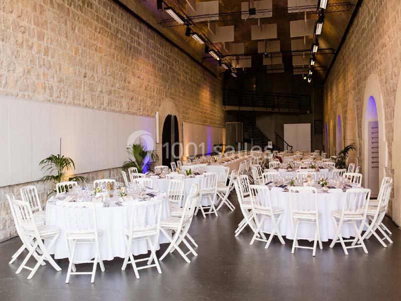 Salle en pierre avec des tables rondes dressées, nappes blanches et chaises alignées pour un événement.