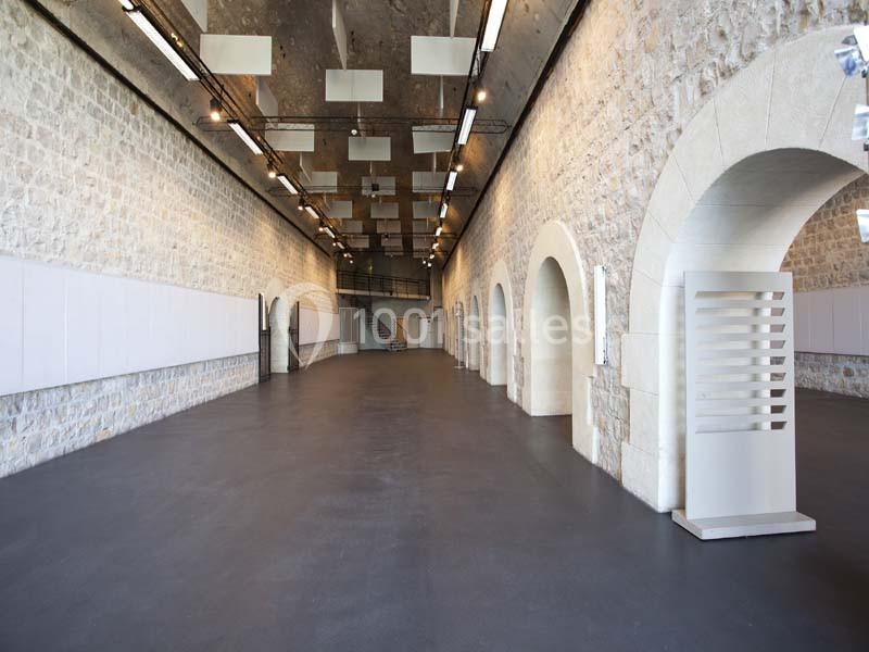 Salle intérieure avec murs en pierre, arches, sol sombre et plafond équipé de luminaires modernes.