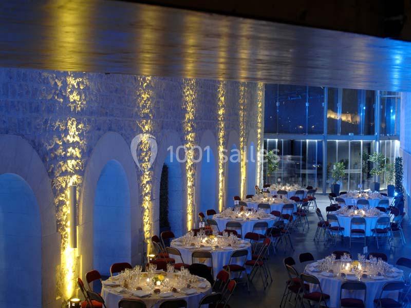Salle de réception avec murs en pierre éclairés, tables rondes dressées et grandes baies vitrées en arrière-plan.