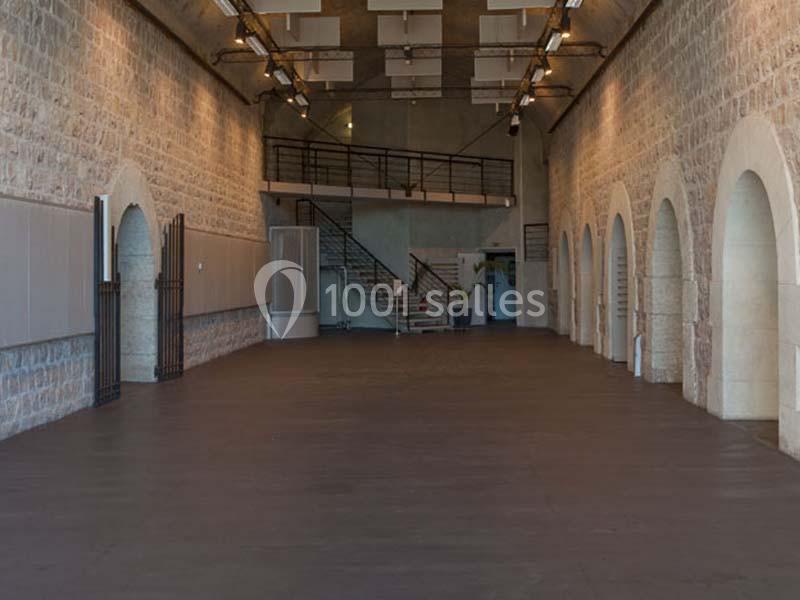 Salle vide avec murs en pierre, arches, sol sombre, escalier métallique et éclairage suspendu.