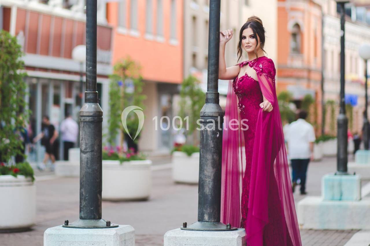 Femme en robe de soirée rouge posant dans une rue bordée de bâtiments et de lampadaires.