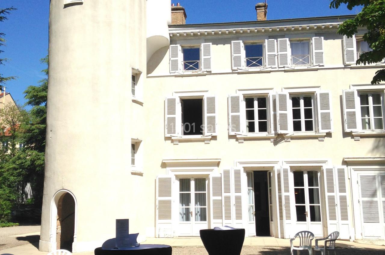 Tour blanche et bâtiment avec volets, tables hautes et chaises disposées dans une cour ensoleillée.