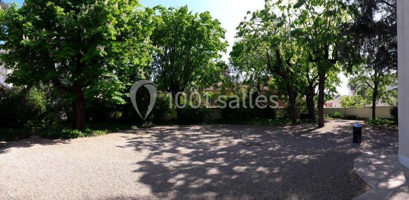 Cour extérieure ombragée avec des arbres feuillus, sol en gravier et quelques bâtiments visibles en arrière-plan.