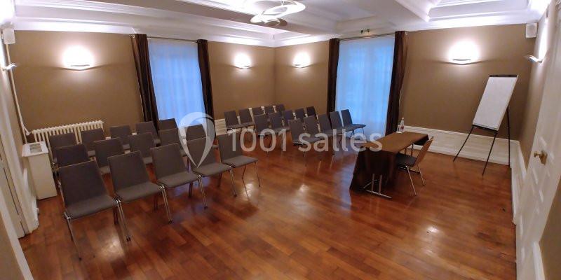 Salle de réunion avec parquet, chaises alignées, table, chevalet de conférence et éclairage mural.