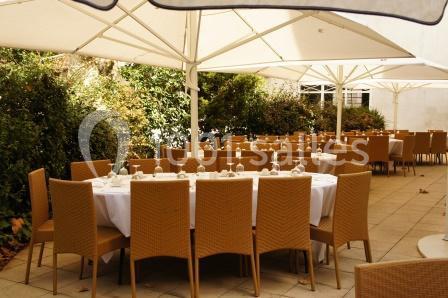 Terrasse aménagée avec des tables rondes dressées, entourées de chaises en osier, sous des parasols blancs.