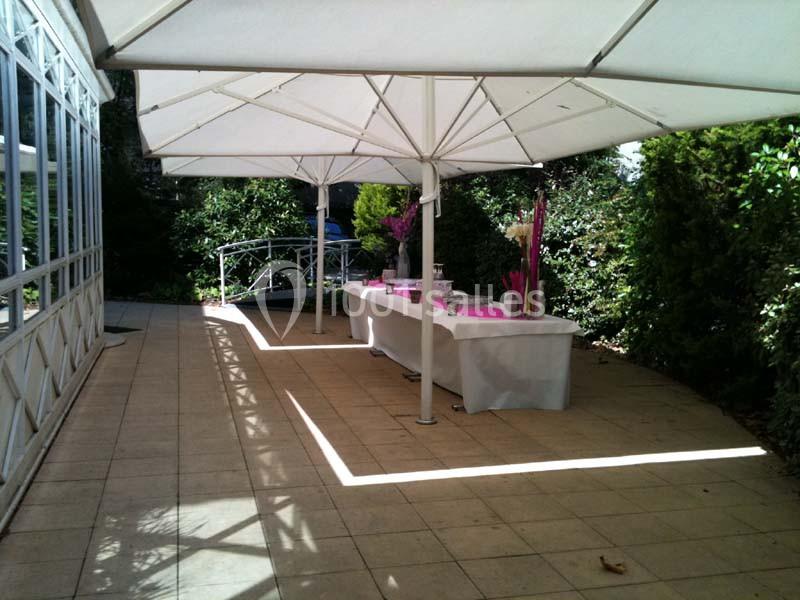 Terrasse avec tables décorées de nappes blanches et ornements roses, abritée par de grands parasols blancs.