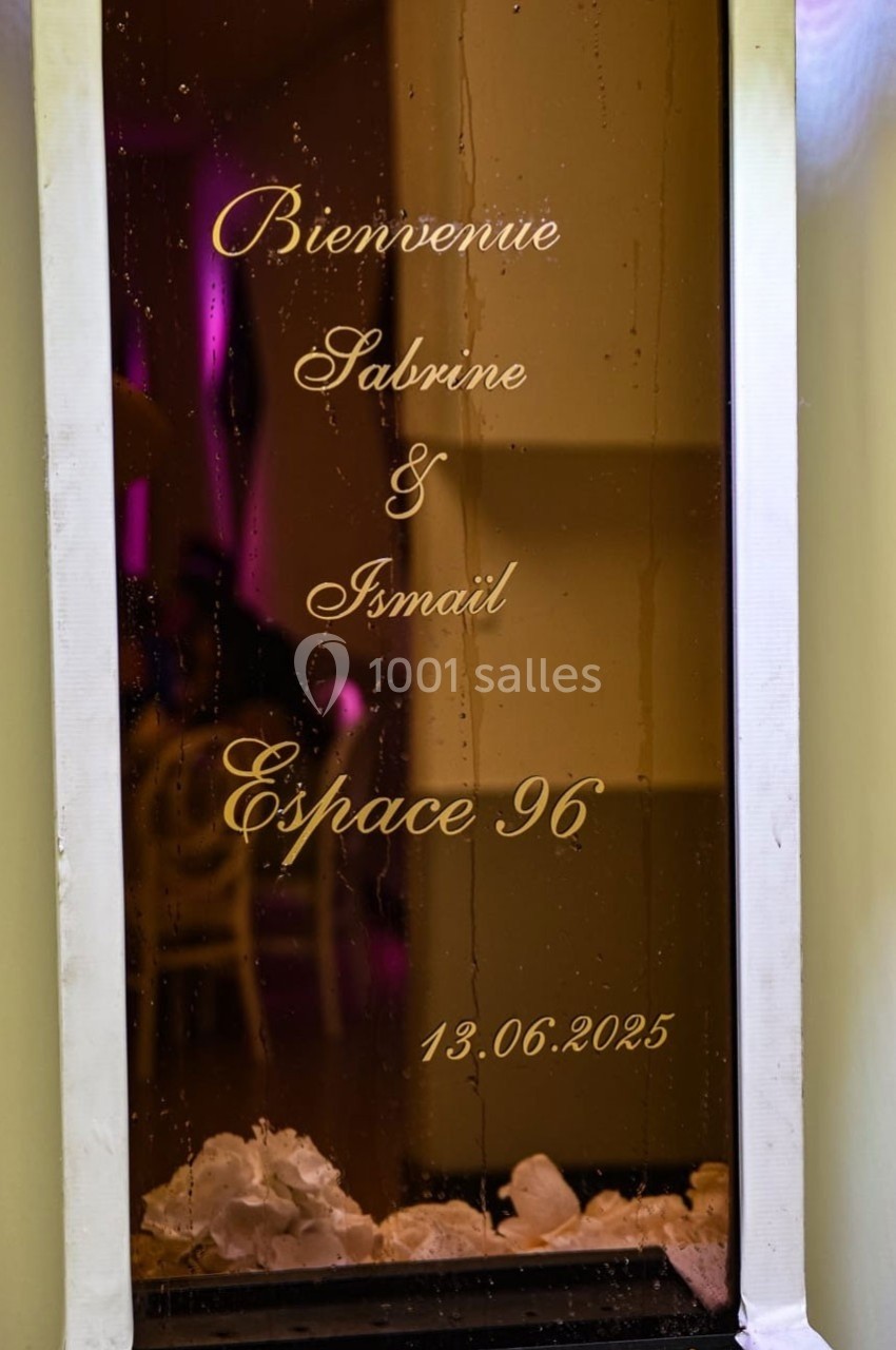 Texte doré sur une vitre indiquant ’Bienvenue Sabrine & Ismail, Espace 96, 13.06.2025’, avec des fleurs blanches en bas.