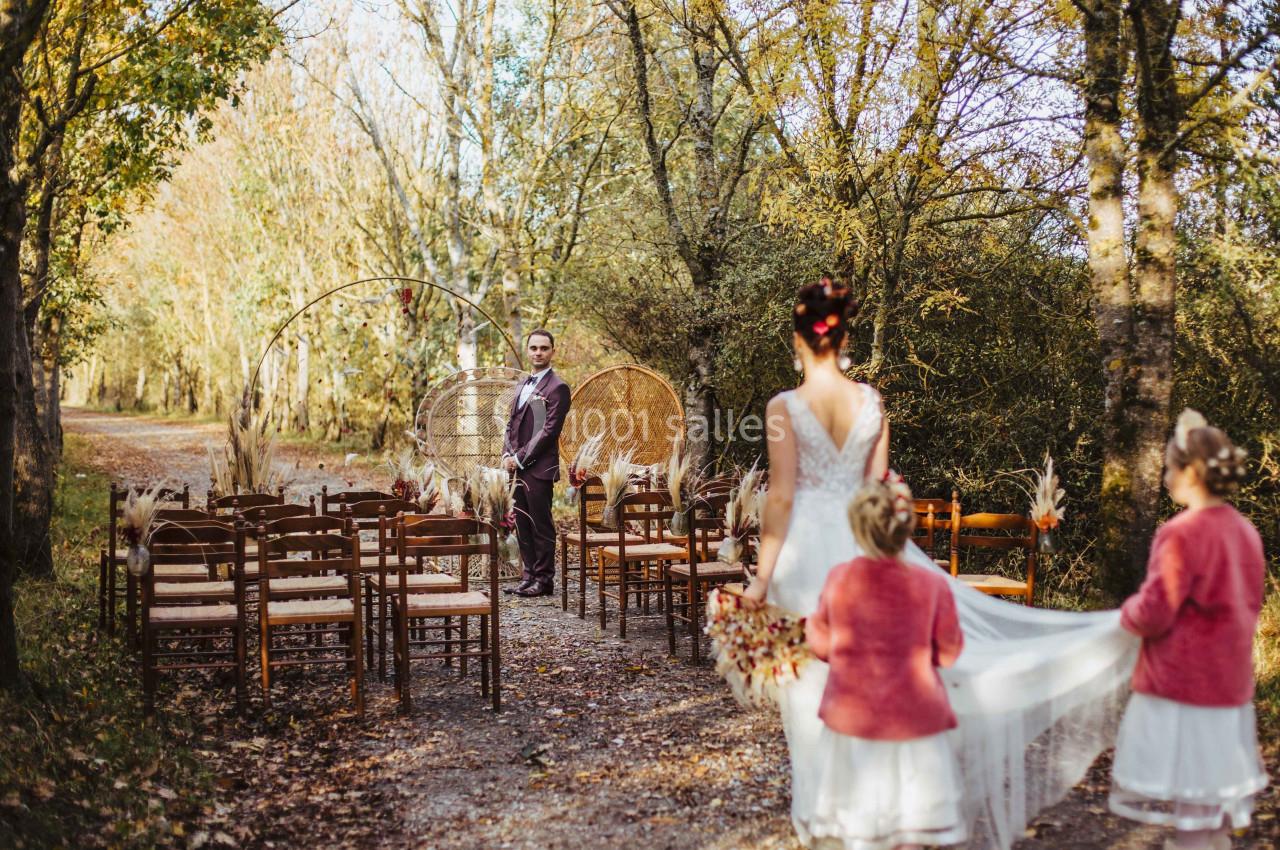 Mariée avançant vers un homme dans une allée extérieure bordée de chaises en bois, entourée d'arbres en automne.