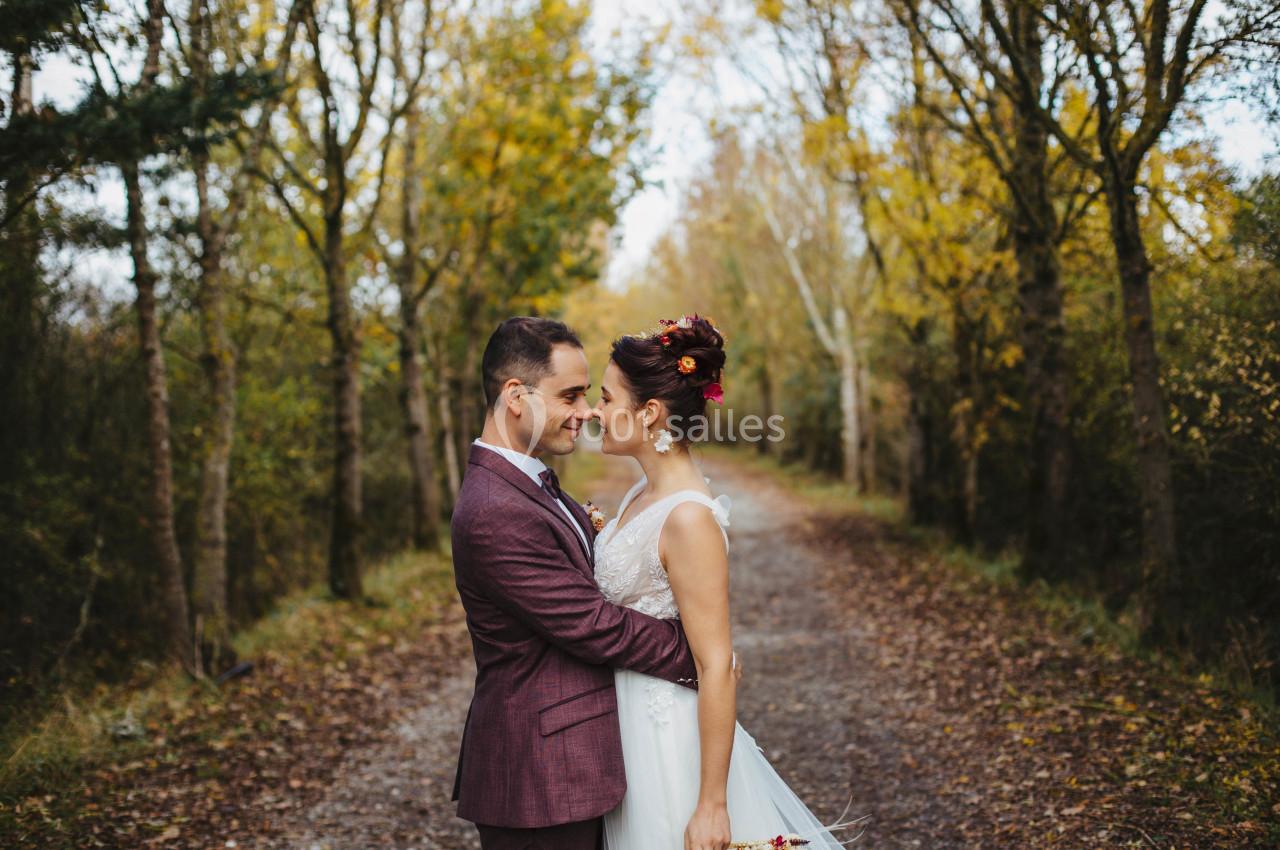 Un couple en tenue de mariage s'enlace sur un chemin bordé d'arbres aux feuilles d'automne.