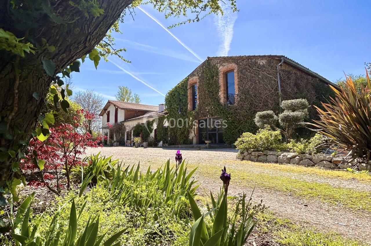 Maison en pierre entourée de végétation, vue depuis un jardin fleuri sous un ciel bleu ensoleillé.
