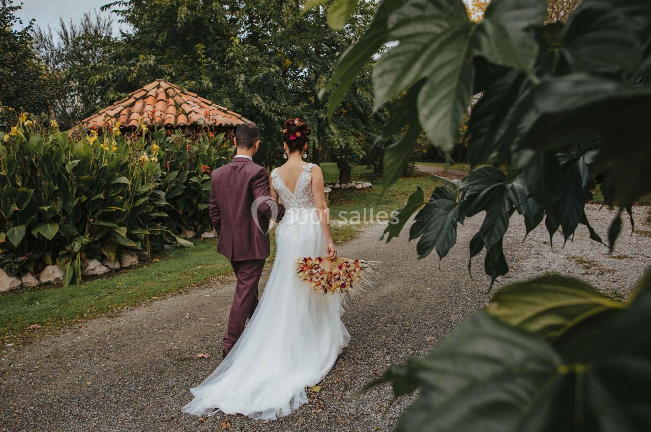 Un couple en tenue de mariage marche sur un chemin bordé de verdure, près d'une petite cabane en pierre.