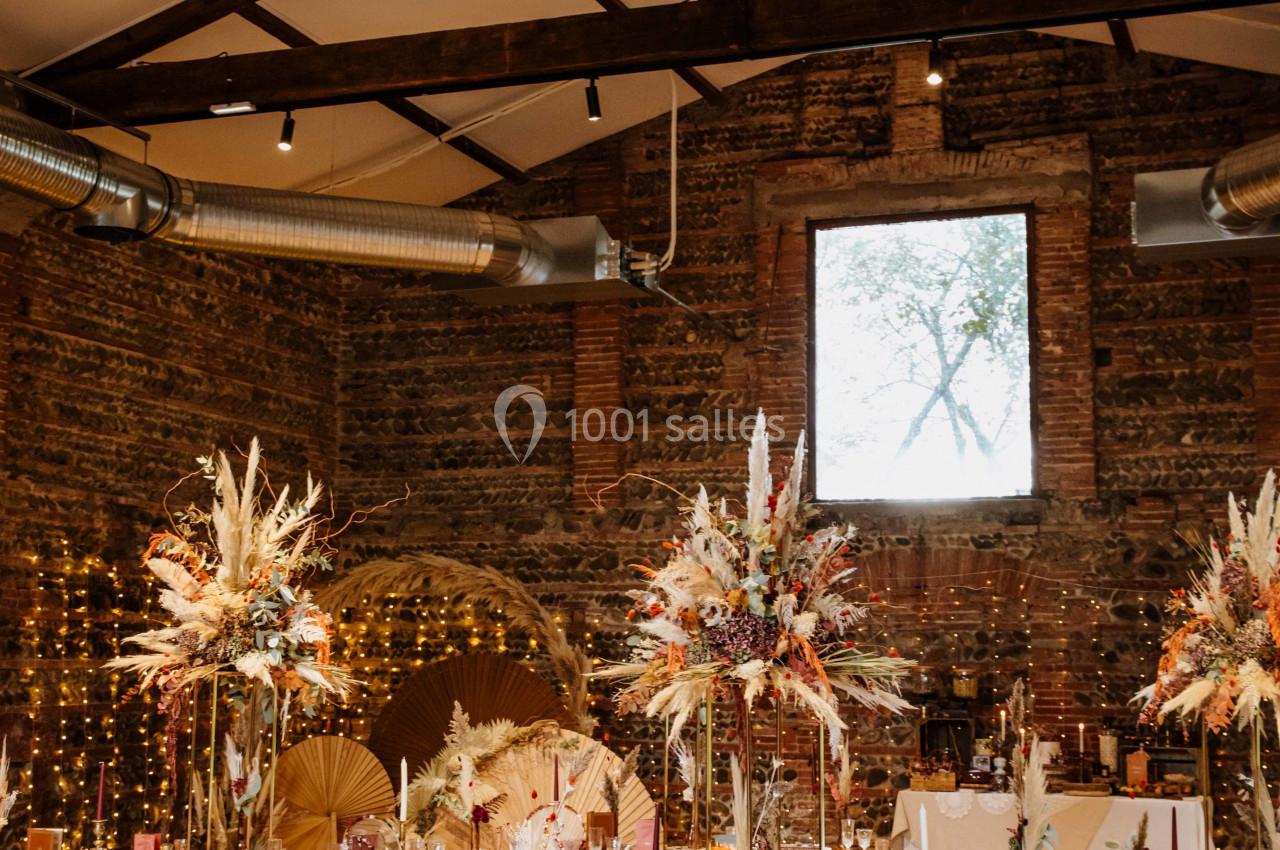 Salle de réception rustique avec tables décorées de nappes blanches, fleurs séchées et guirlandes lumineuses.