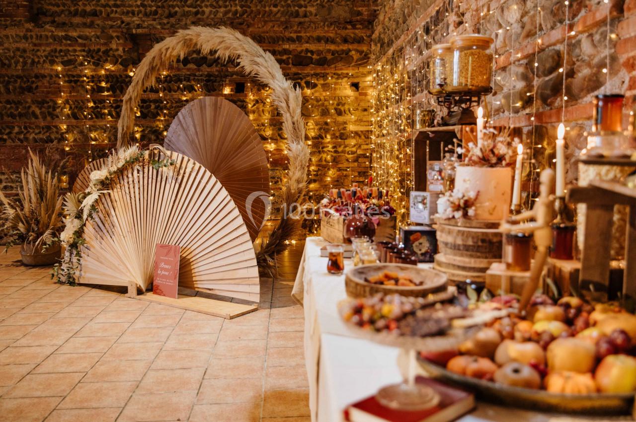 Décoration de mariage avec arche en pampas, éventails en bois, guirlandes lumineuses et table garnie de desserts.
