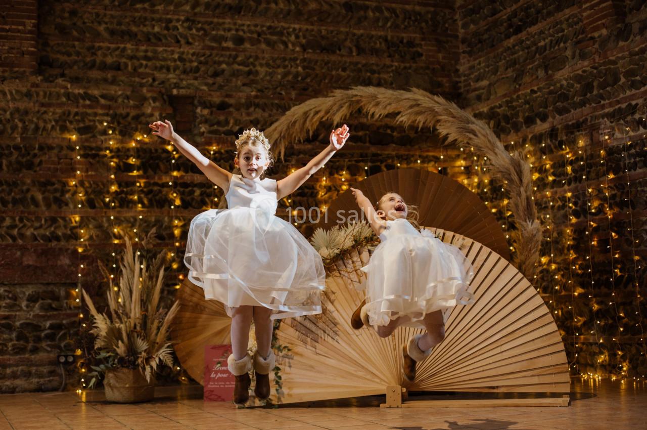 Deux enfants en robes blanches sautent joyeusement devant une décoration lumineuse et végétale dans une salle en briques.