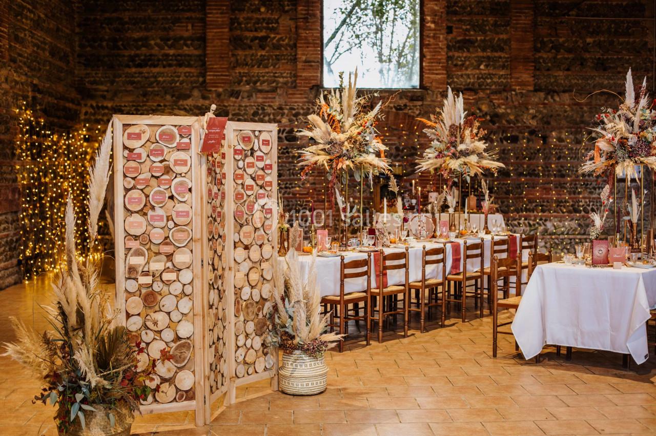 Salle de réception décorée avec des tables dressées, des compositions florales et un panneau en bois pour les invités.
