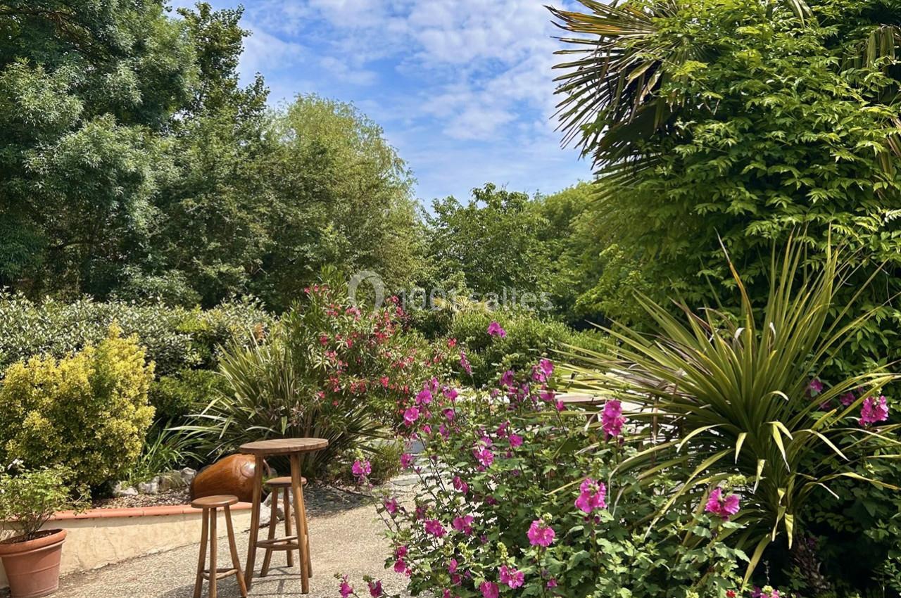 Jardin verdoyant avec fleurs roses, arbustes et palmiers sous un ciel partiellement nuageux.