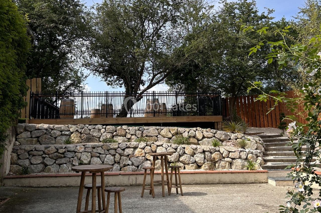 Terrasse surélevée avec tables et chaises, entourée d'arbres et de pierres, vue depuis une cour avec tabourets en bois.