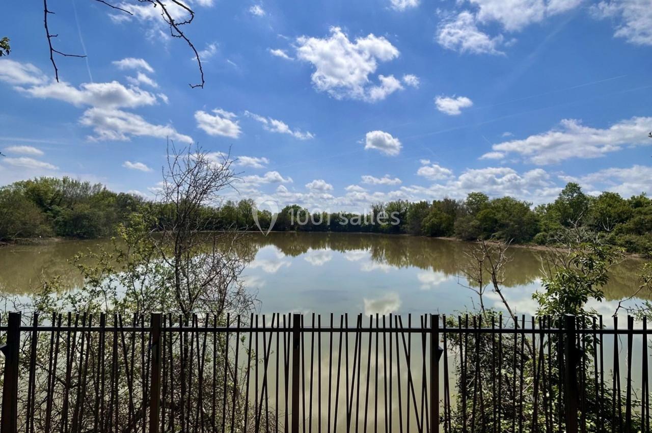 Vue d'un lac calme entouré d'arbres sous un ciel partiellement nuageux, avec une clôture au premier plan.