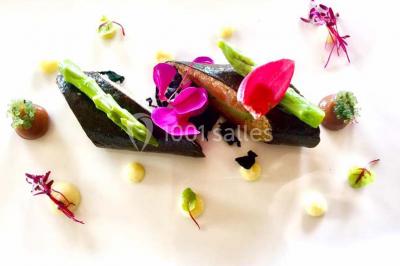 Salle de réception avec tables rondes dressées, nappes blanches, assiettes dorées et décoration florale centrale.