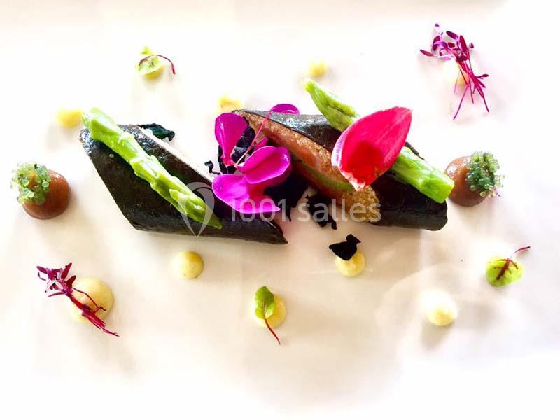 Assiette gastronomique décorée avec des légumes, fleurs comestibles et touches de sauce sur un fond blanc.