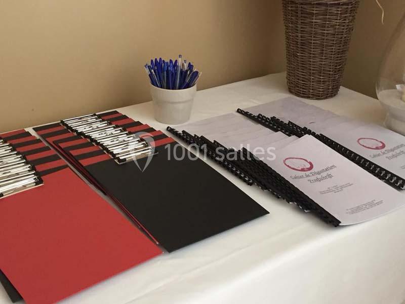 Des porte-blocs rouges et noirs, des stylos bleus dans un pot et des documents reliés posés sur une table blanche.