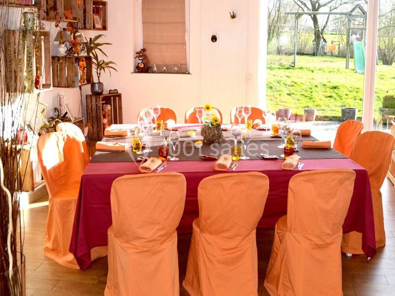 Table dressée avec nappes colorées, chaises houssées orange, vaisselle et décoration florale, vue sur jardin en arrière-plan.