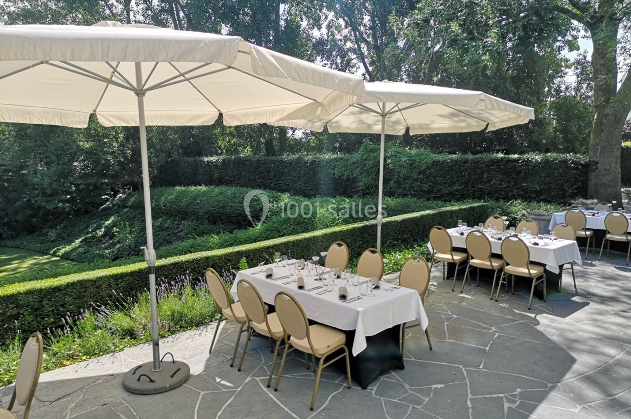 Tables dressées sous des parasols blancs sur une terrasse en pierre, entourée de verdure et d'arbustes taillés.