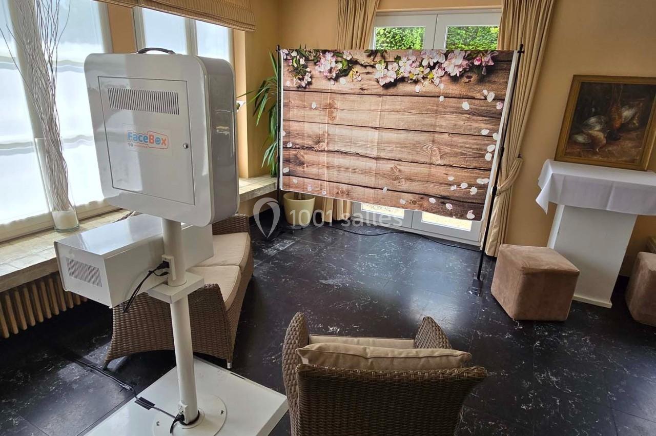 Cabine photo installée dans une pièce lumineuse avec un fond en bois décoré de fleurs et des fauteuils en osier.