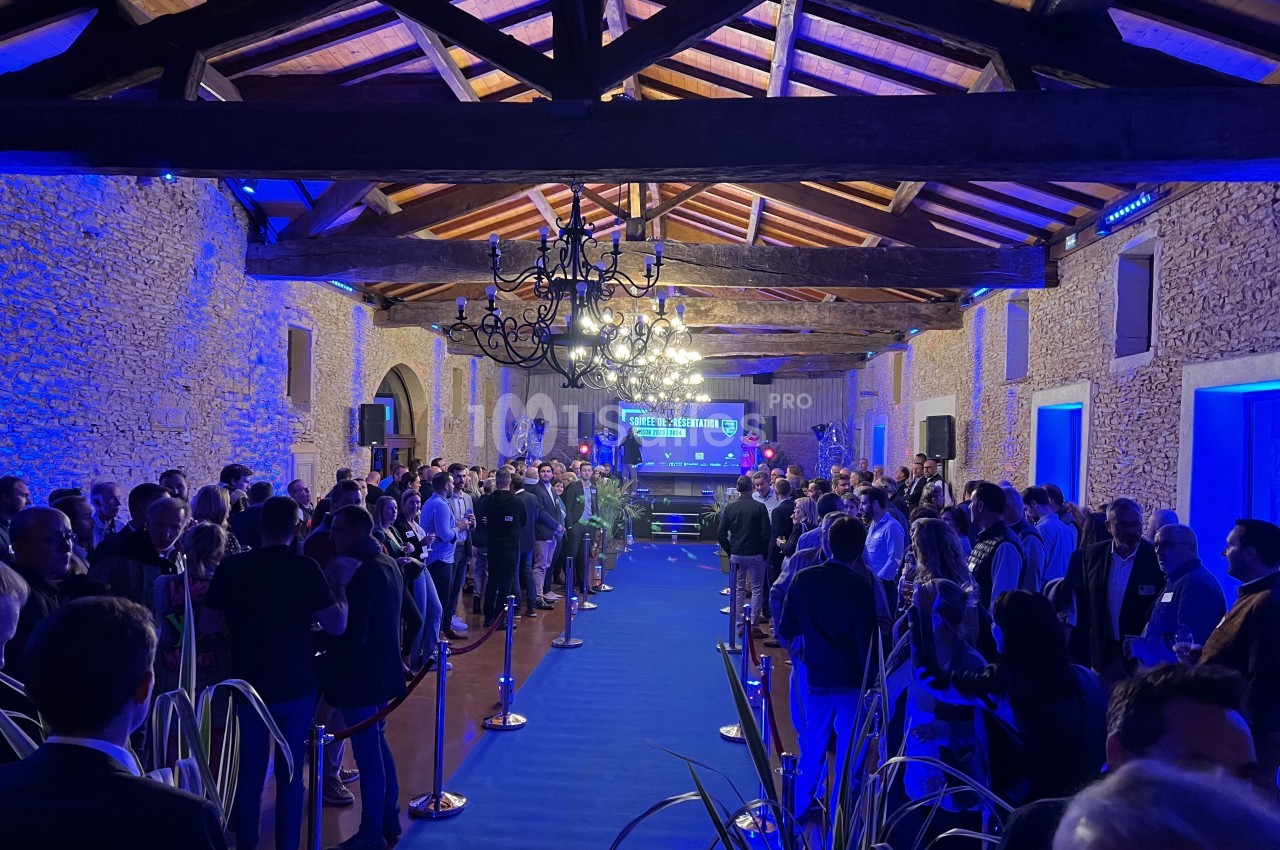 Salle en pierre avec plafond en bois, éclairée en bleu, remplie de personnes lors d'un événement avec une scène au fond.