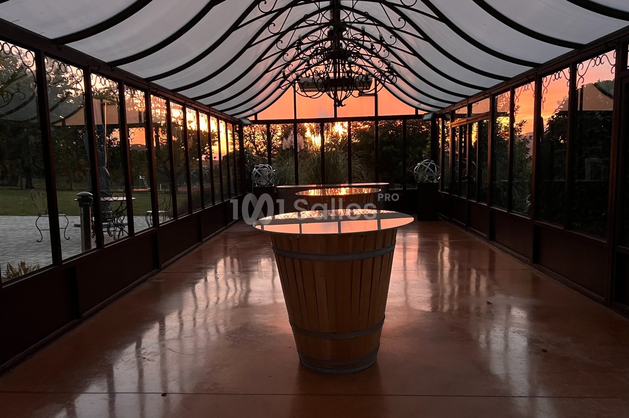 Intérieur d'une serre vitrée au coucher du soleil, avec une table en tonneau au centre et un sol brillant.