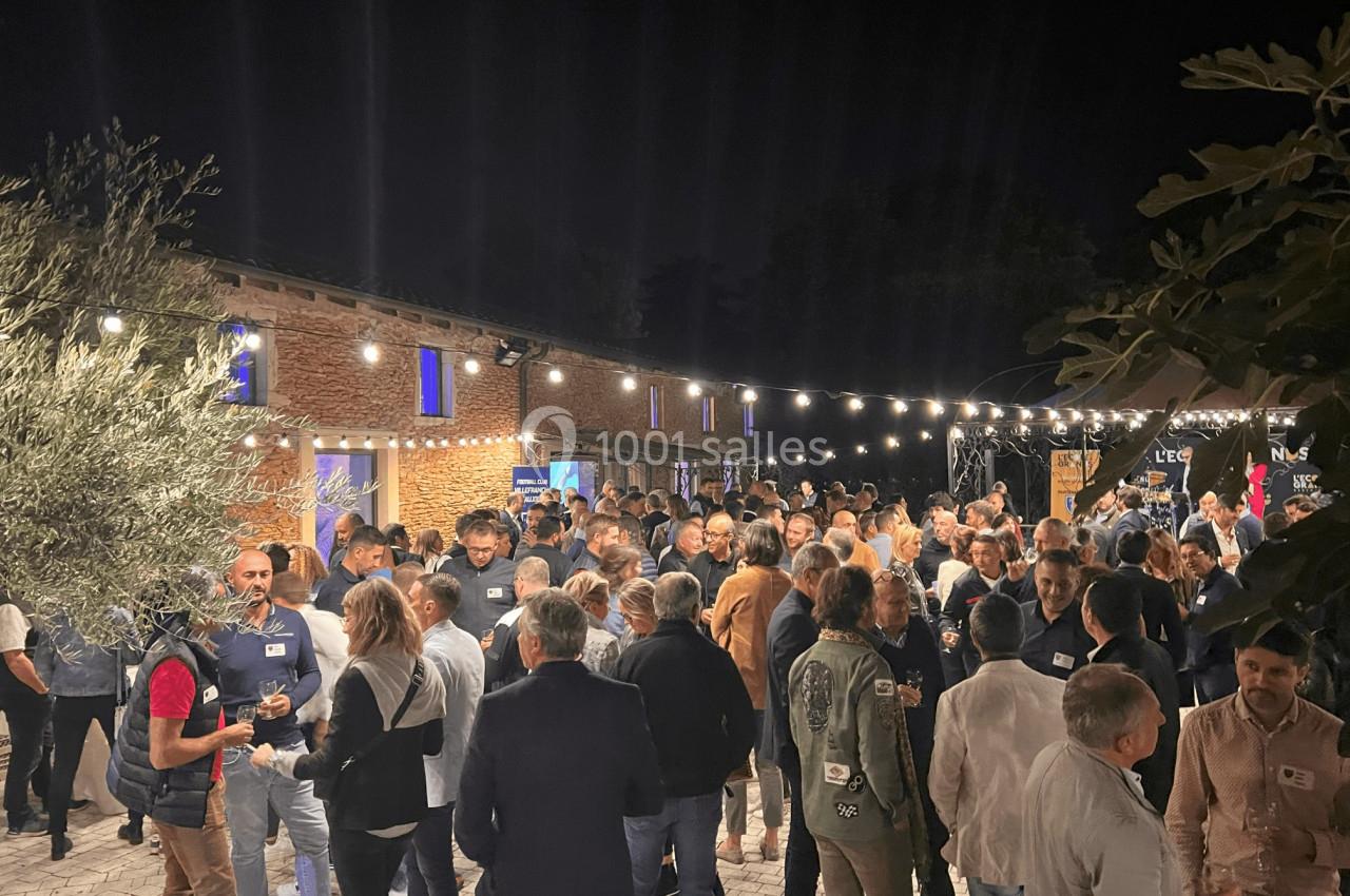 Groupe de personnes rassemblées en soirée dans une cour éclairée par des guirlandes lumineuses, devant des bâtiments en…
