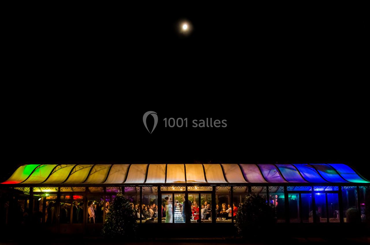 Vue nocturne d'une salle vitrée éclairée par des lumières colorées sous un ciel sombre avec une pleine lune.