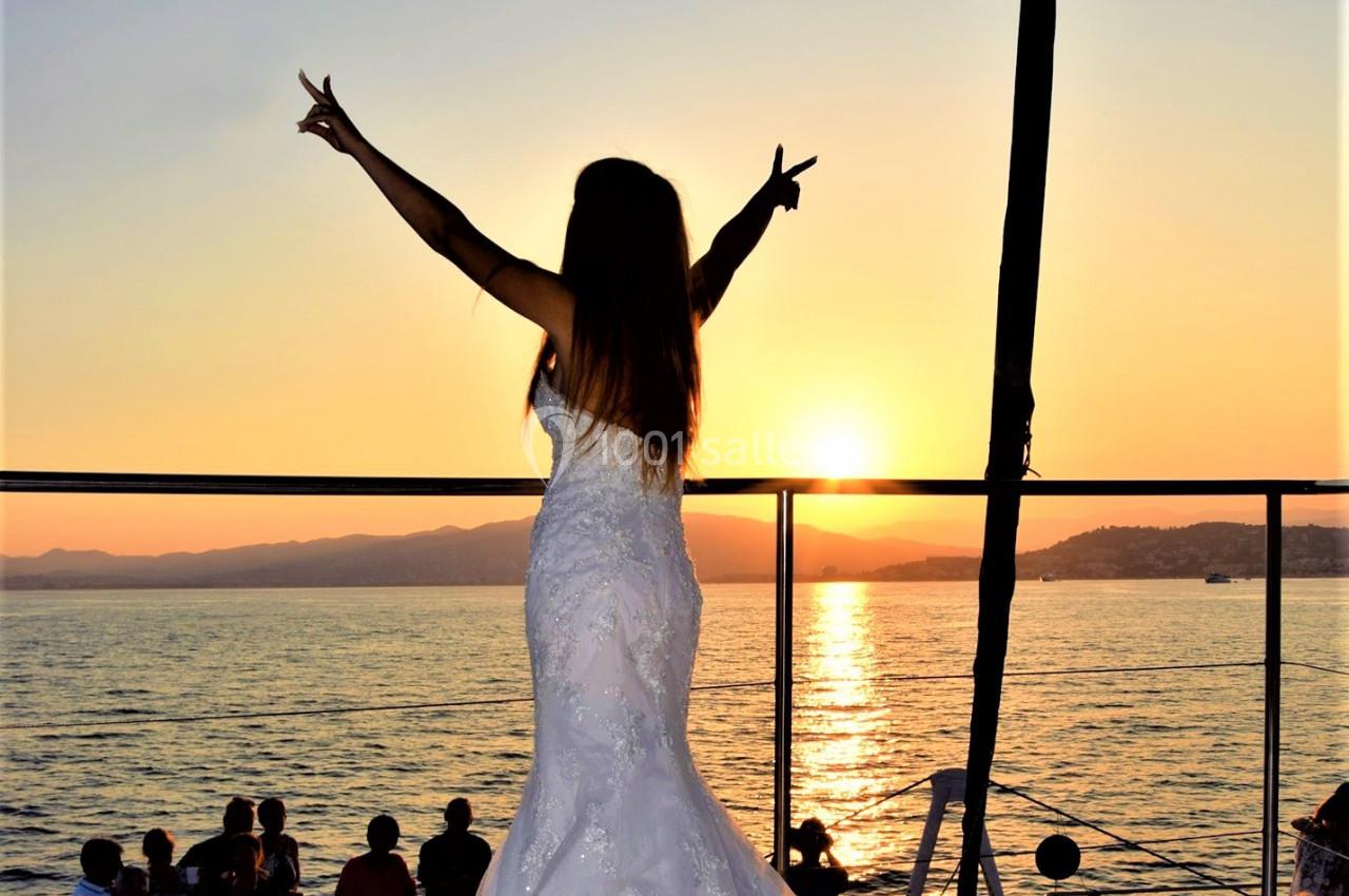 Femme en robe de mariée levant les bras face à un coucher de soleil sur un bateau, entourée de spectateurs.