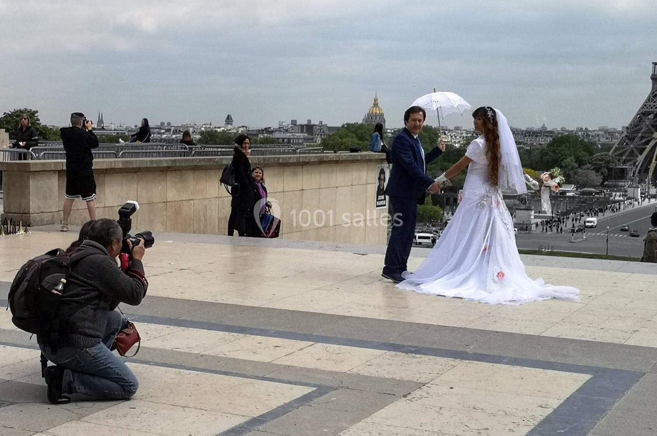 Un couple en tenue de mariage pose devant un photographe sur une esplanade avec vue sur la tour Eiffel.