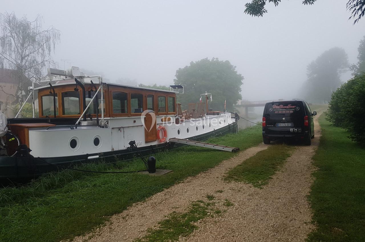 Bateau amarré le long d'un chemin de gravier dans un paysage brumeux avec un véhicule stationné à proximité.