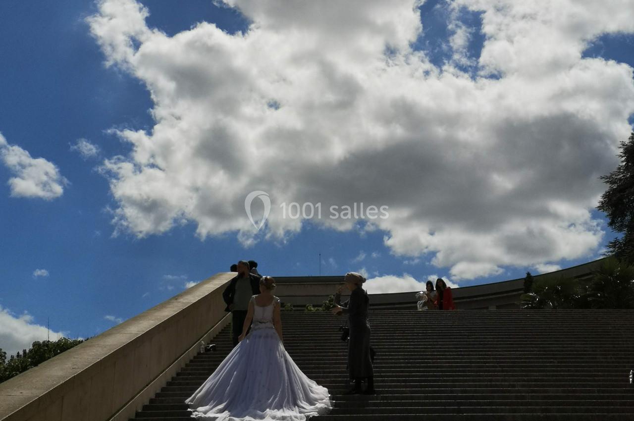 Un couple en tenue de mariage pose sur un large escalier extérieur sous un ciel partiellement nuageux.