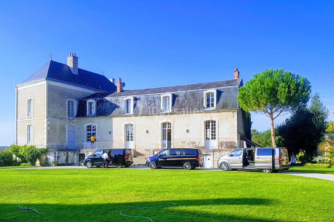 Façade d'un grand bâtiment ancien entouré de verdure, avec plusieurs véhicules stationnés devant.