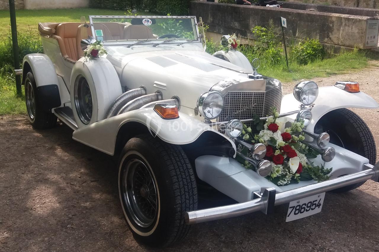 Voiture ancienne blanche décorée de fleurs, stationnée en extérieur sur un chemin ensoleillé.
