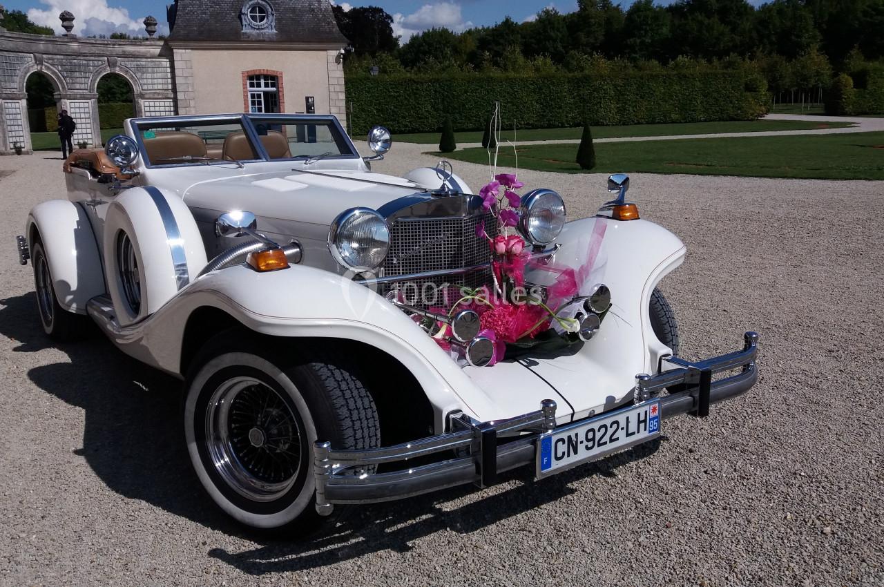 Voiture ancienne blanche décorée de fleurs roses, stationnée devant un bâtiment historique et un jardin.