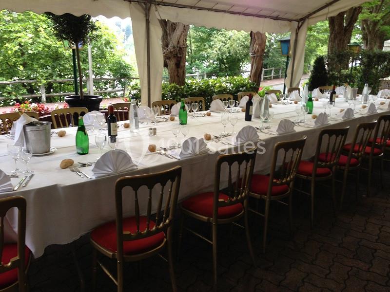 Table dressée sous une tente, avec nappes blanches, serviettes pliées, bouteilles et chaises à assise rouge.